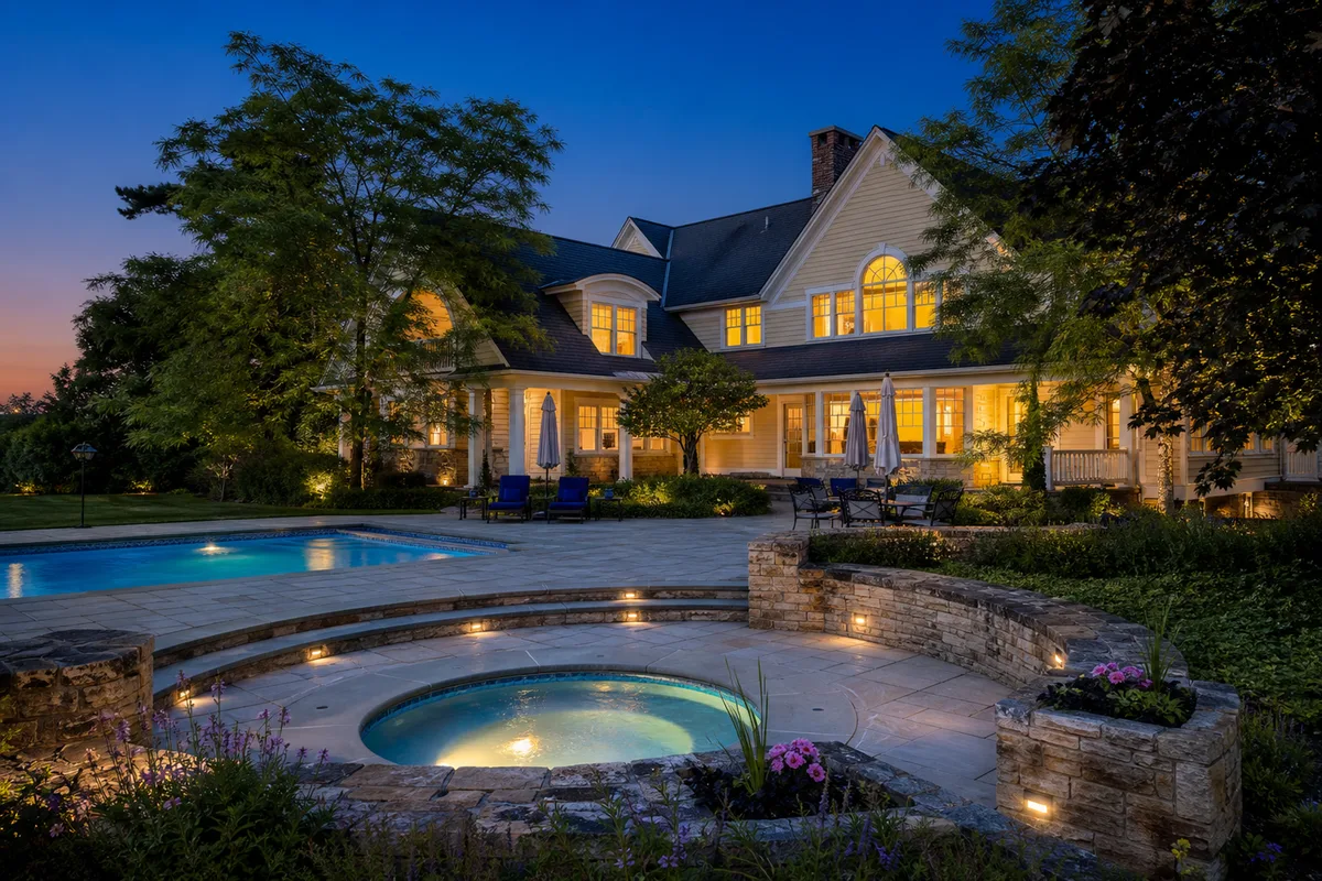 Rear exterior twilight view of the estate, showcasing the glowing warm windows overlooking the stone-paved patio and private swimming pool.